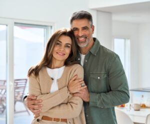 Smiling affectionate loving middle aged couple, happy romantic mature older Latin man and woman in their 50s hugging looking at camera standing together in modern kitchen at home. Portrait.